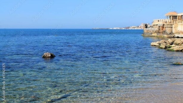Un bâtiment situé sur la côte, avec une eau d’un bleu limpide au premier plan et une côte rocheuse.