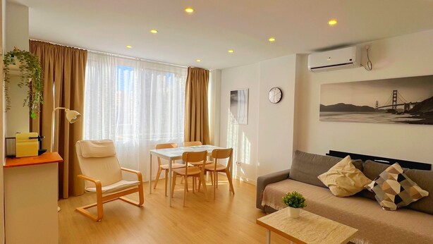 Una sala moderna con un sofá, una mesa de comedor con sillas, una pequeña mesa de centro y una zona de cocina con un refrigerador y una planta.