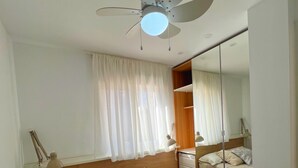 Un dormitorio con cama, ventilador de techo, ventana con cortinas, lámpara de noche y espejo.