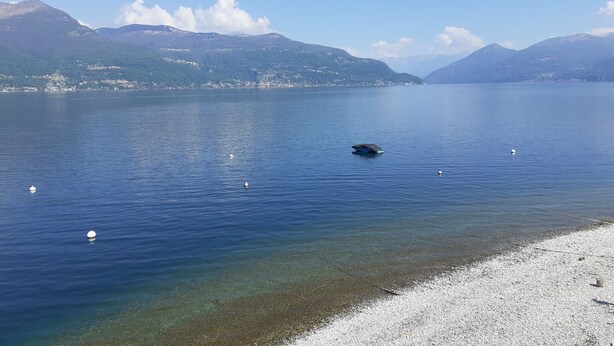 Ein ruhiger See mit klarem Wasser, kiesigem Ufer und Bergen im Hintergrund.