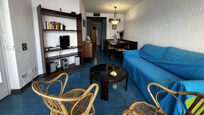 Ein Wohnzimmer mit blauem Sofa, Korbstühlen, einem Couchtisch, einem Bücherregal und einer Essecke mit Tisch und Stühlen.