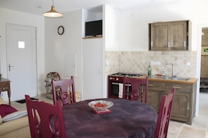 Een eethoek met een tafel, stoelen, een televisie boven de tafel, een keuken met kasten en een fornuis, en een klok aan de muur.