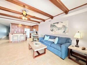 A living room with a blue sofa, a coffee table, and a lamp. There is a kitchen in the background with wooden cabinets and a ceiling fan.