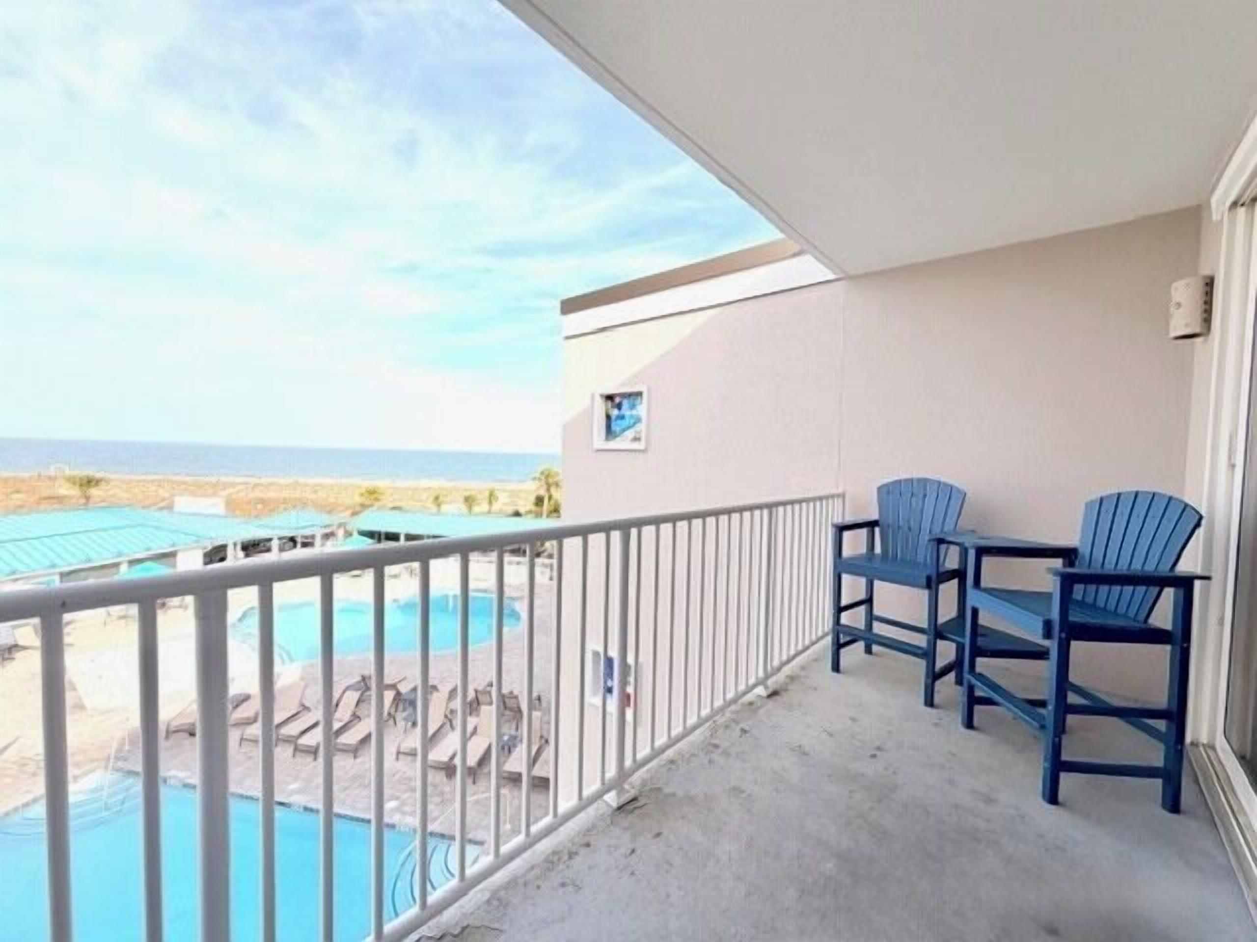 A balcony with a view of a pool and beach, two blue chairs, and a small picture frame.