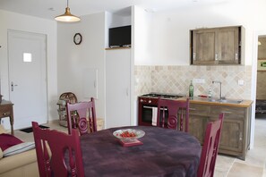 En matplats med ett bord, stolar, en tv ovanför bordet, ett kök med skåp och en spis samt en klocka på väggen.