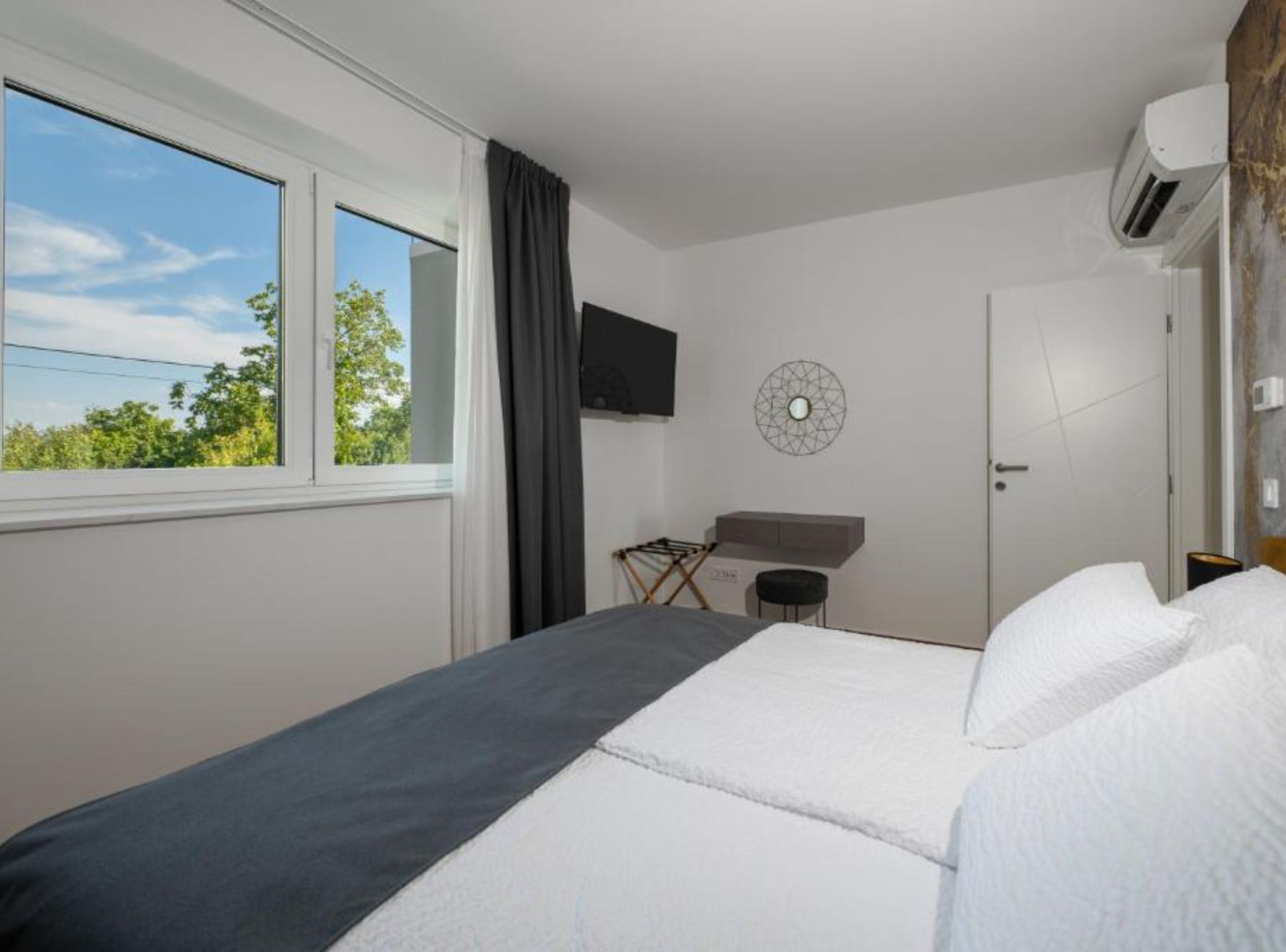 A hotel room with a bed, a TV mounted on the wall, a window with curtains, and a view of trees outside.