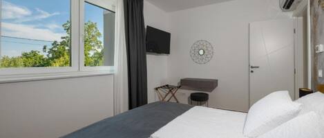 A hotel room with a bed, a TV mounted on the wall, a window with curtains, and a view of trees outside.
