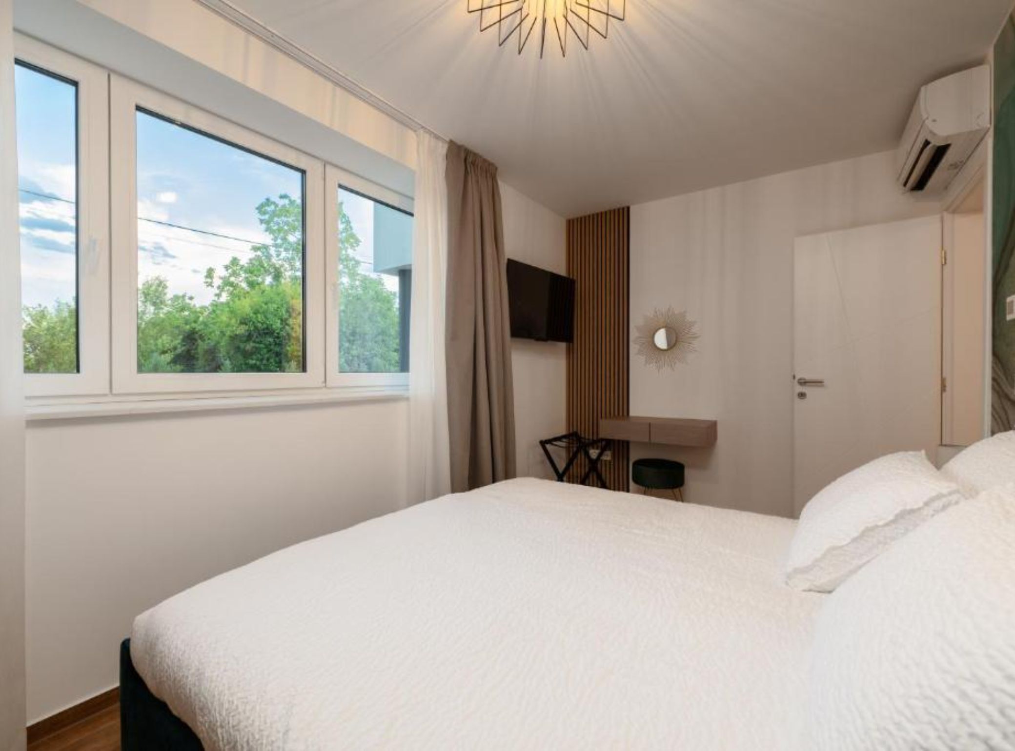 Een slaapkamer met een groot bed, een aan de muur bevestigde televisie, een raam met gordijnen en een deur.