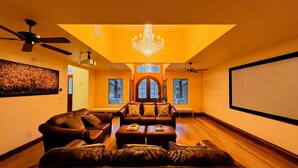 A spacious living room with a large sectional sofa, a coffee table, and a chandelier. There are two windows with arched tops and a ceiling fan.