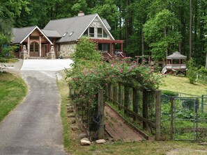 Une maison en pierre à deux étages, dotée d’une cheminée en pierre, d’un porche en bois et d’un belvédère, située dans une zone boisée.