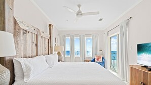 Quarto espaçoso com uma cama grande, ventilador de teto, televisão e vista para o mar através de grandes janelas.