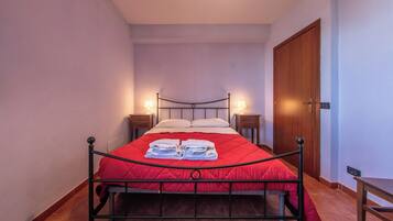 Un lit simple avec une couverture rouge, deux tables de chevet avec des lampes, une porte en bois et un petit banc.