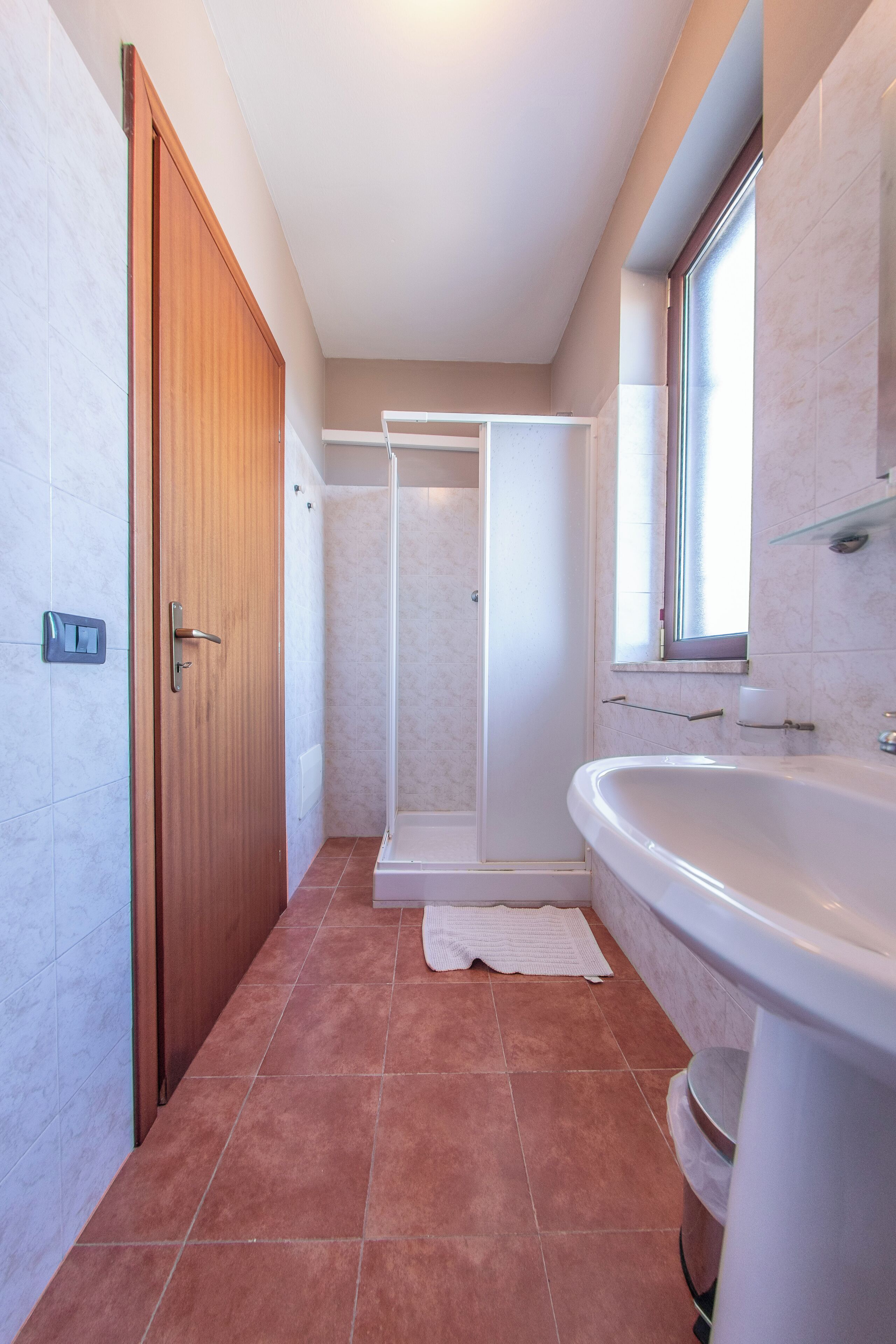 A bathroom with a wooden door, a shower, a sink, and a window.