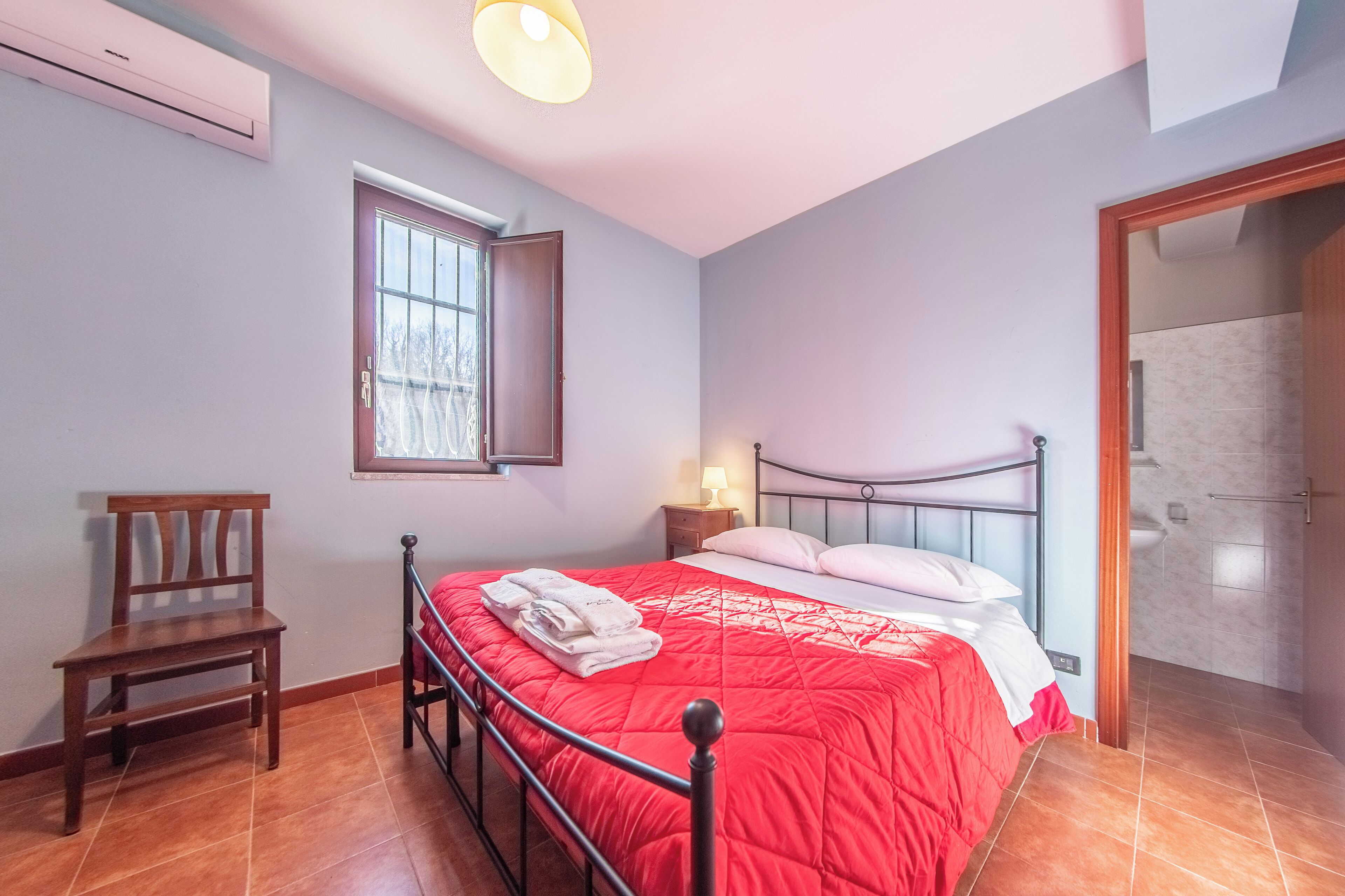 A bedroom with a bed, a chair, a nightstand with a lamp, and a window.
