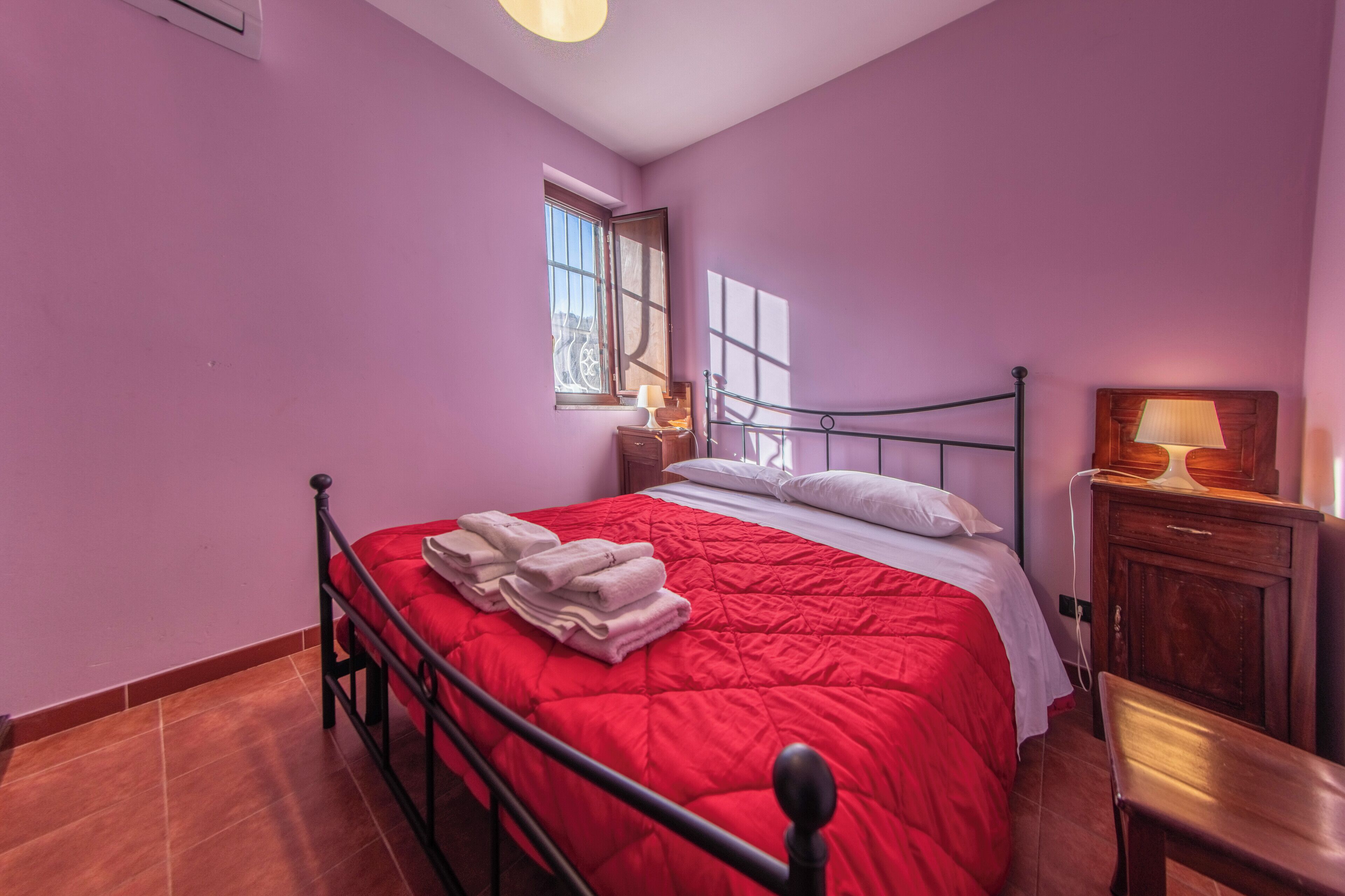Une chambre avec une couvre-lit rouge, une table de chevet en bois avec une lampe, une fenêtre donnant sur un beau paysage et une chaise en bois.