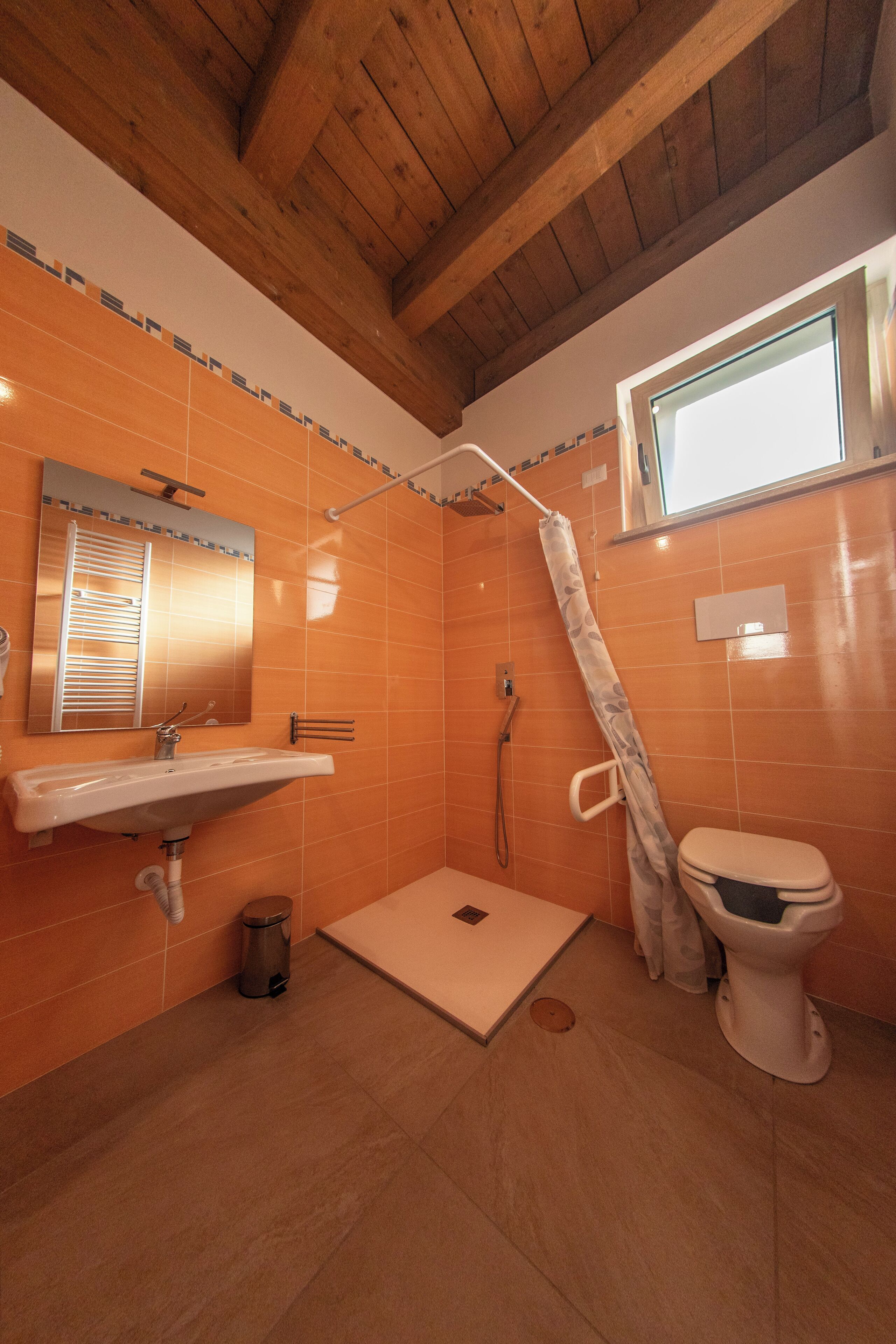 Et badeværelse med orange fliser, et træloft, en hvid vask, et toilet og et bruseområde.