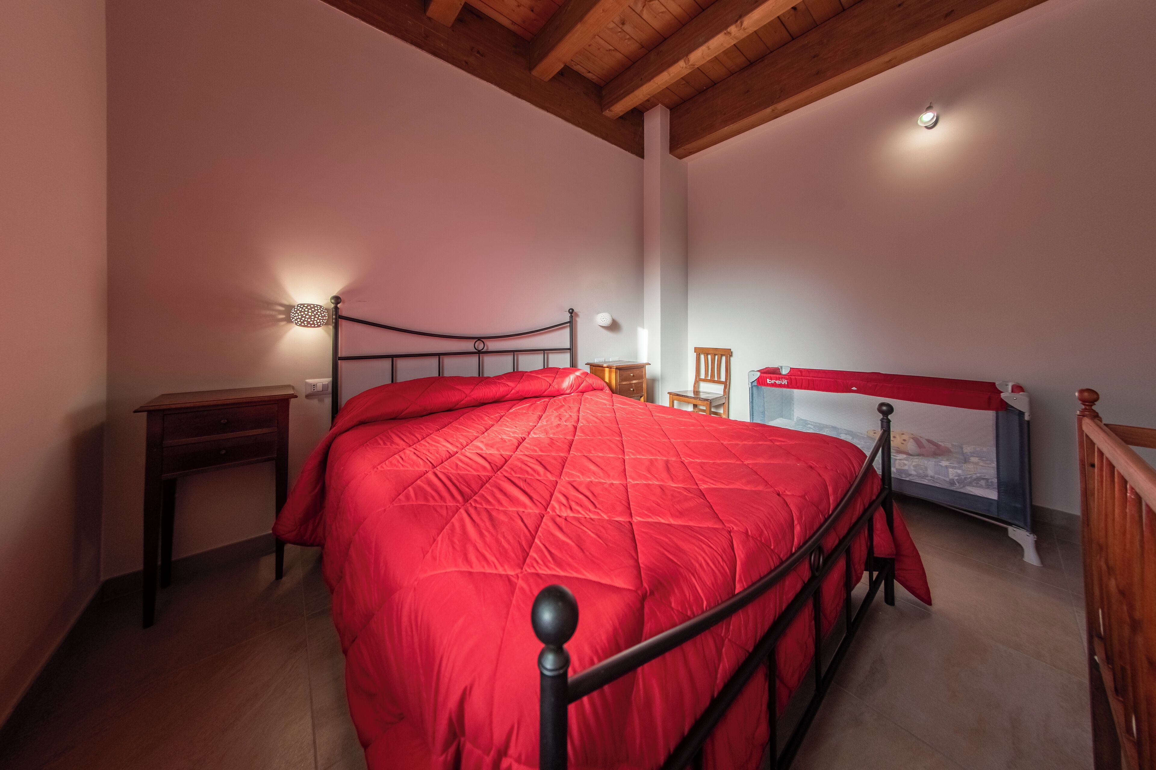 Un dormitorio con una colcha roja, cabecera de madera, mesita de noche, una pequeña mesa y estante montado en la pared.