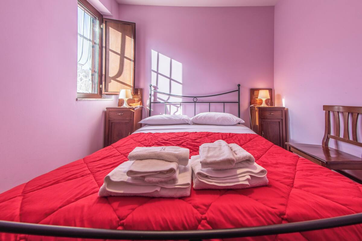 Un lit bien fait, recouvert d’une couverture rouge, deux tables de chevet avec des lampes et une fenêtre laissant entrer la lumière du soleil.