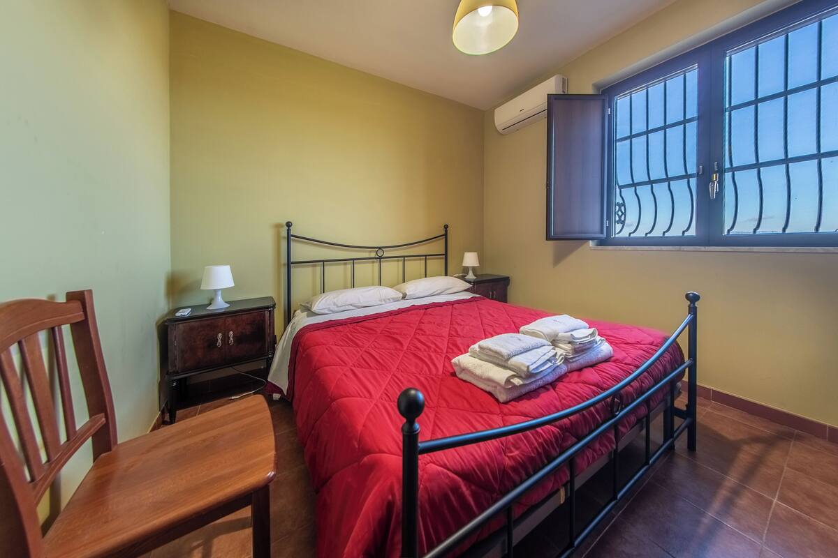 Une chambre à coucher avec un lit, une chaise, une table de chevet, une lampe et une fenêtre avec des barreaux.