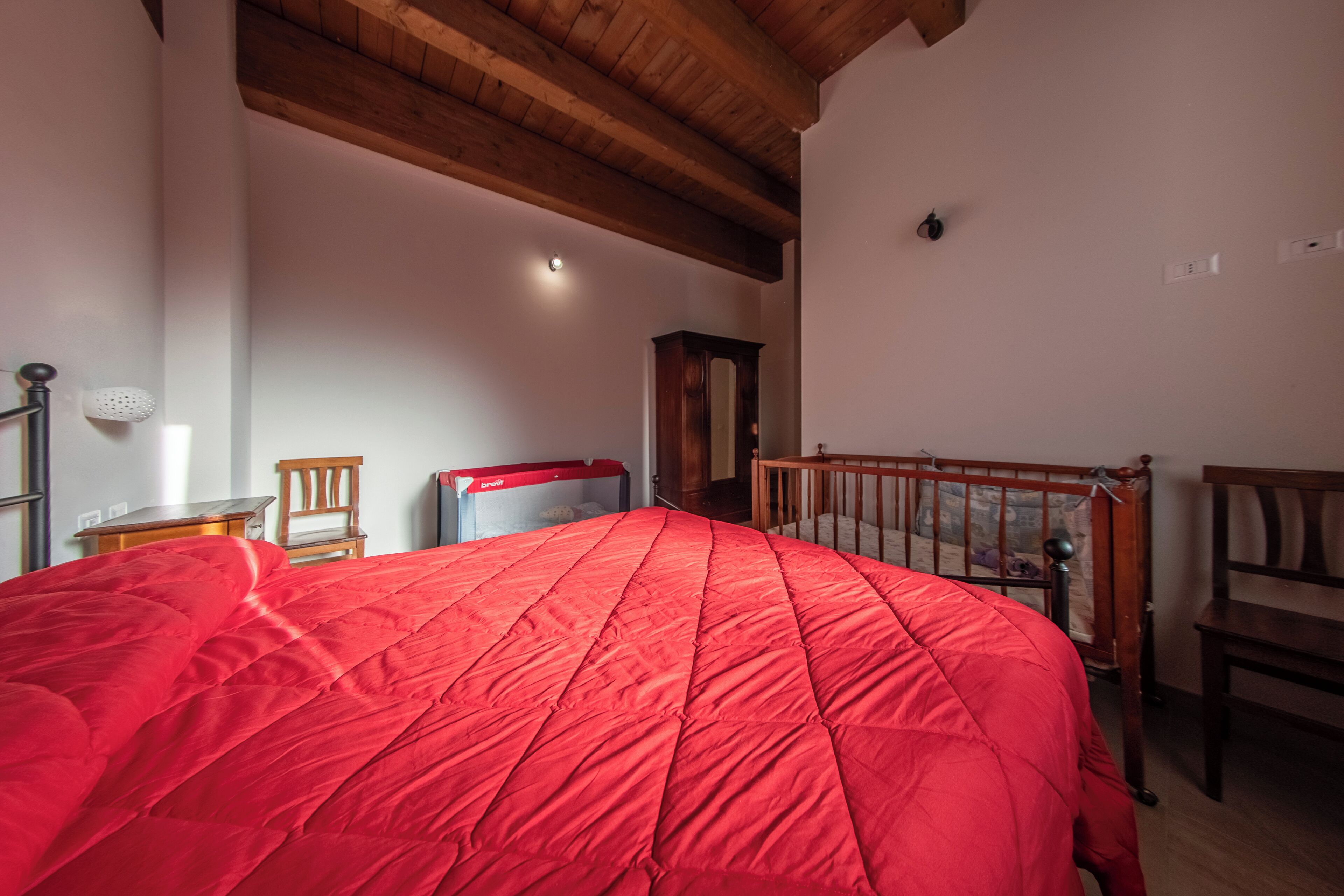 Une chambre avec un couvre-lit rouge, des poutres en bois au plafond, une chaise en bois, une table de chevet et une petite table.