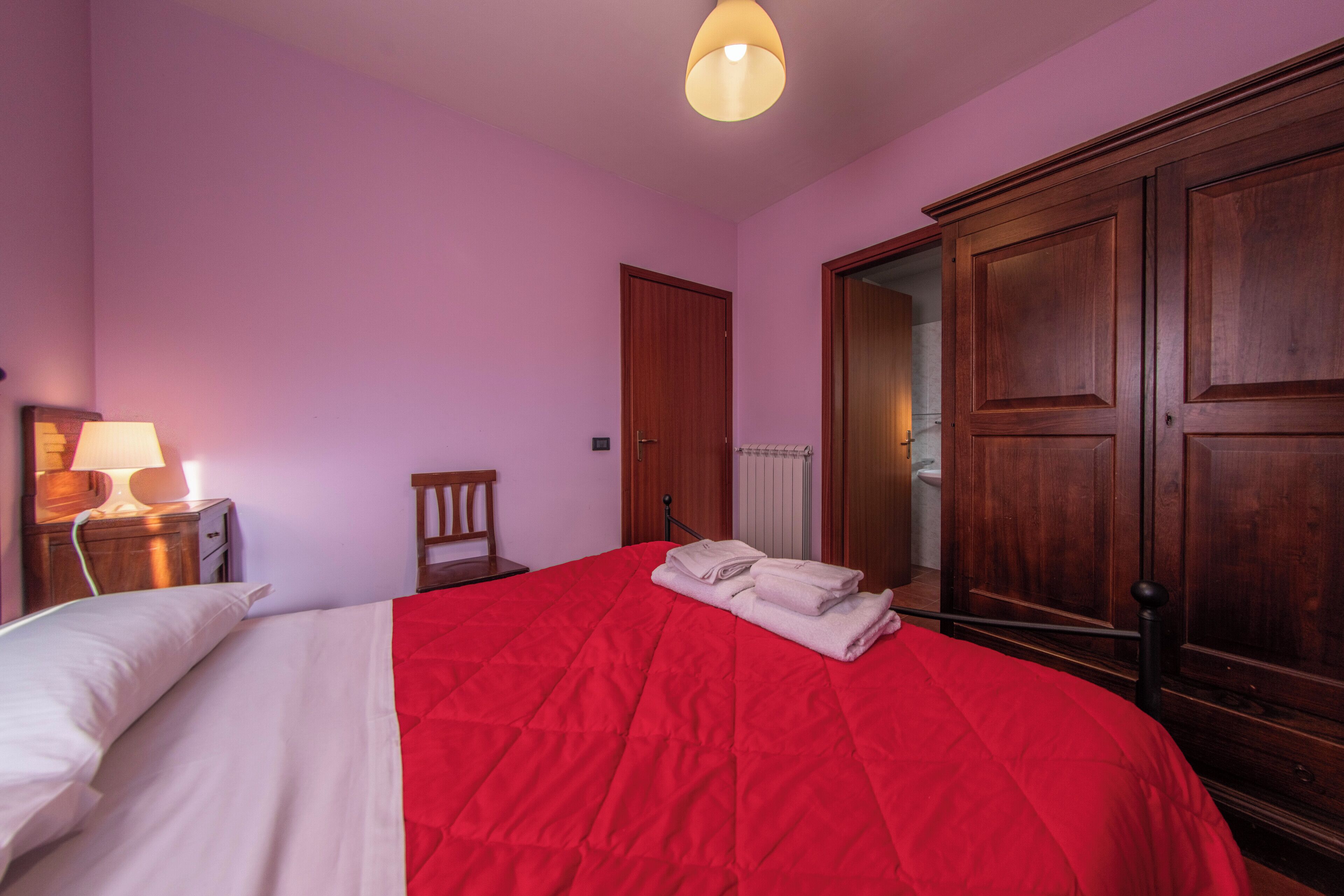 A bedroom with a red bedspread, a wooden wardrobe, a nightstand with a lamp, and a door leading to a bathroom.