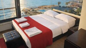 A hotel room with a bed, a red blanket, and a view of the sea and buildings.
