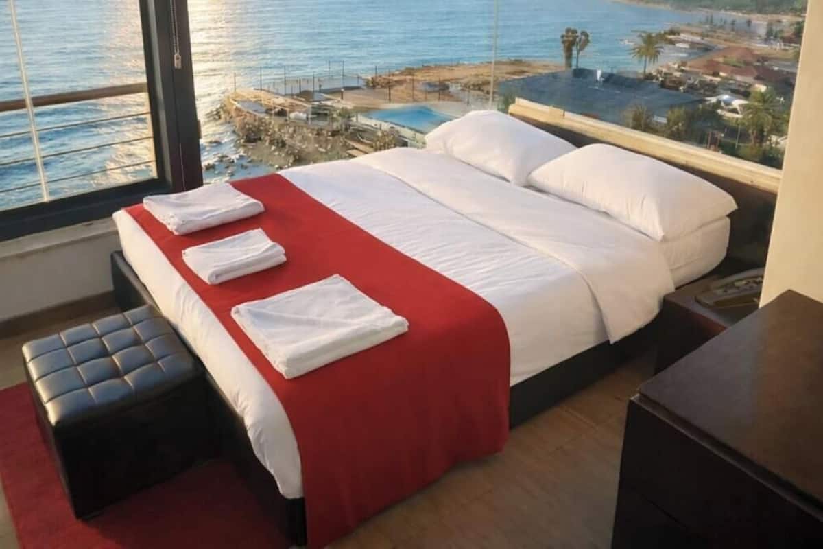Une chambre d’hôtel avec un lit, une couverture rouge et une vue sur la mer et les bâtiments.