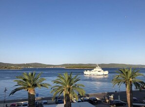 Blick auf die Küste mit einem weißen Boot namens 'IADIGOLINA' auf dem Wasser, Palmen und geparkten Autos am Ufer.