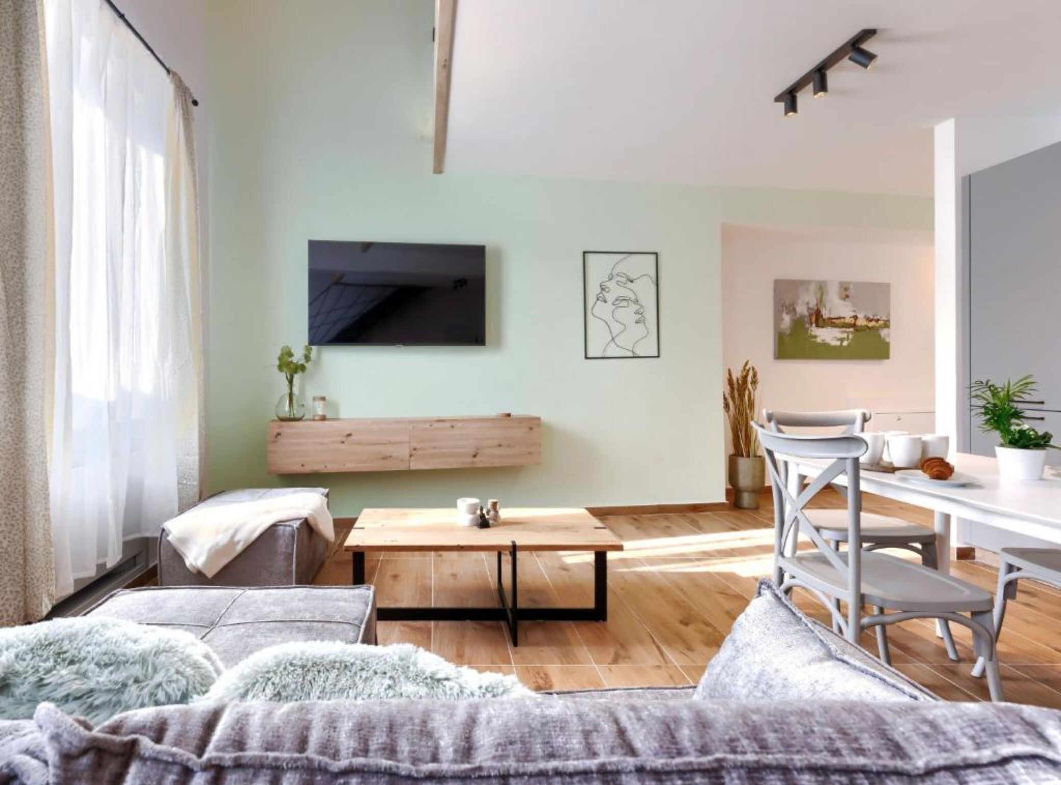 A modern living room with a sofa, a coffee table, a TV mounted on the wall, and a dining area with chairs and a table.