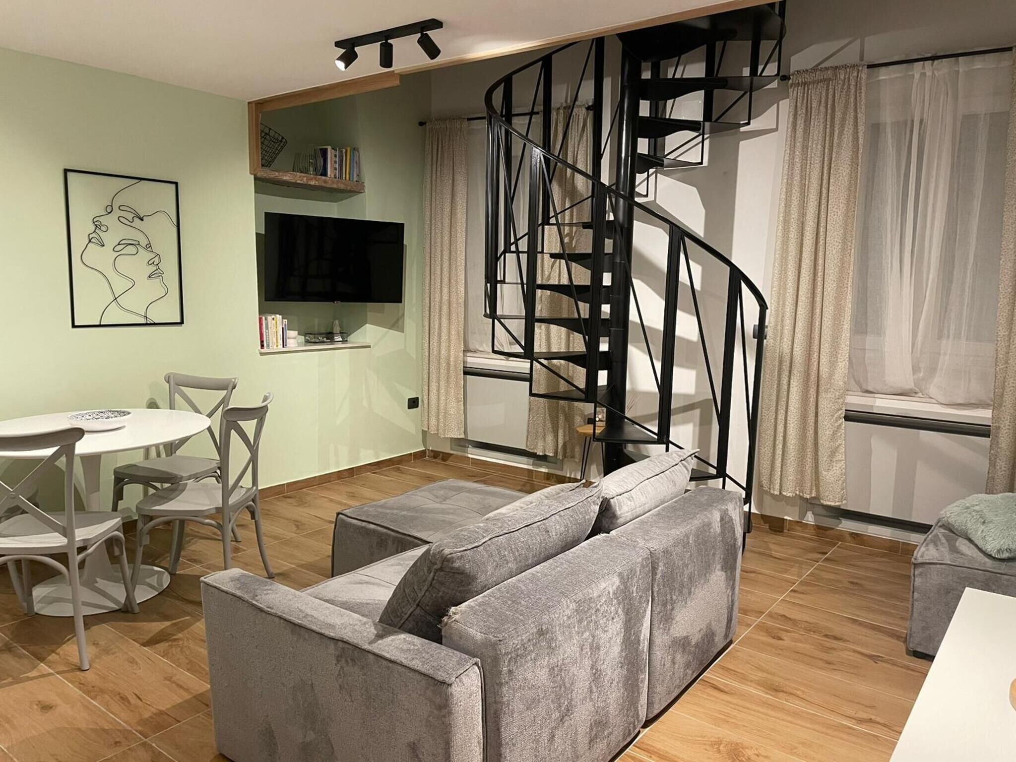 A modern living room with a grey sofa, a dining table with chairs, a TV, a staircase, and a bookshelf.