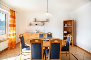 A dining area with a round table, six chairs, a hanging light fixture, wooden shelves, and a bookshelf.