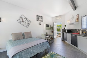 Une chambre moderne avec un lit, des tables de chevet et un petit bureau avec une chaise. On peut apercevoir une kitchenette équipée d’un four à micro-ondes, d’une cuisinière et d’un évier.