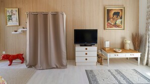 Quarto com televisão sobre suporte de madeira, cómoda branca com gavetas, estátua de um cão vermelho, cortina bege e um quadro na parede.