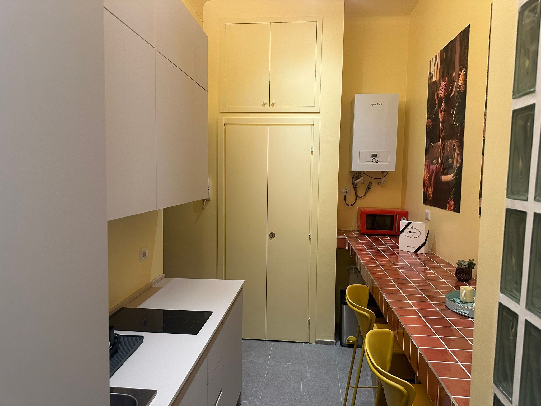 A compact kitchen with a white countertop, yellow chairs, a red microwave, and a white water heater.