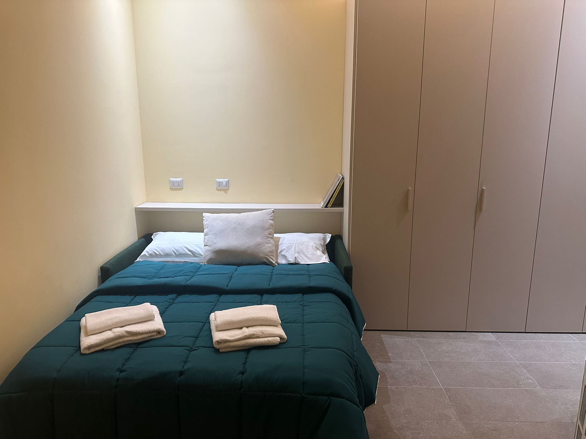 Una cama pequeña y ordenada con una colcha azul verdoso y dos almohadas blancas, un par de toallas dobladas sobre la cama, una mesita de noche blanca con un libro y una lámpara, y un armario cerrado.