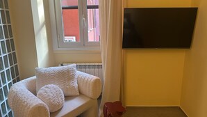 A room with a white cushioned bench, a flat-screen TV mounted on the wall, and a window with curtains. - Atelier 12 (Milan)