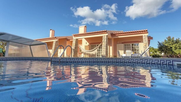 Une piscine, un ciel d’un bleu limpide et un bâtiment doté d’un toit en tuiles et de piliers en pierre.