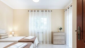 Une chambre d’hôtel avec deux lits, une commode en bois, une fenêtre avec des rideaux légers et une porte dotée d’une poignée.