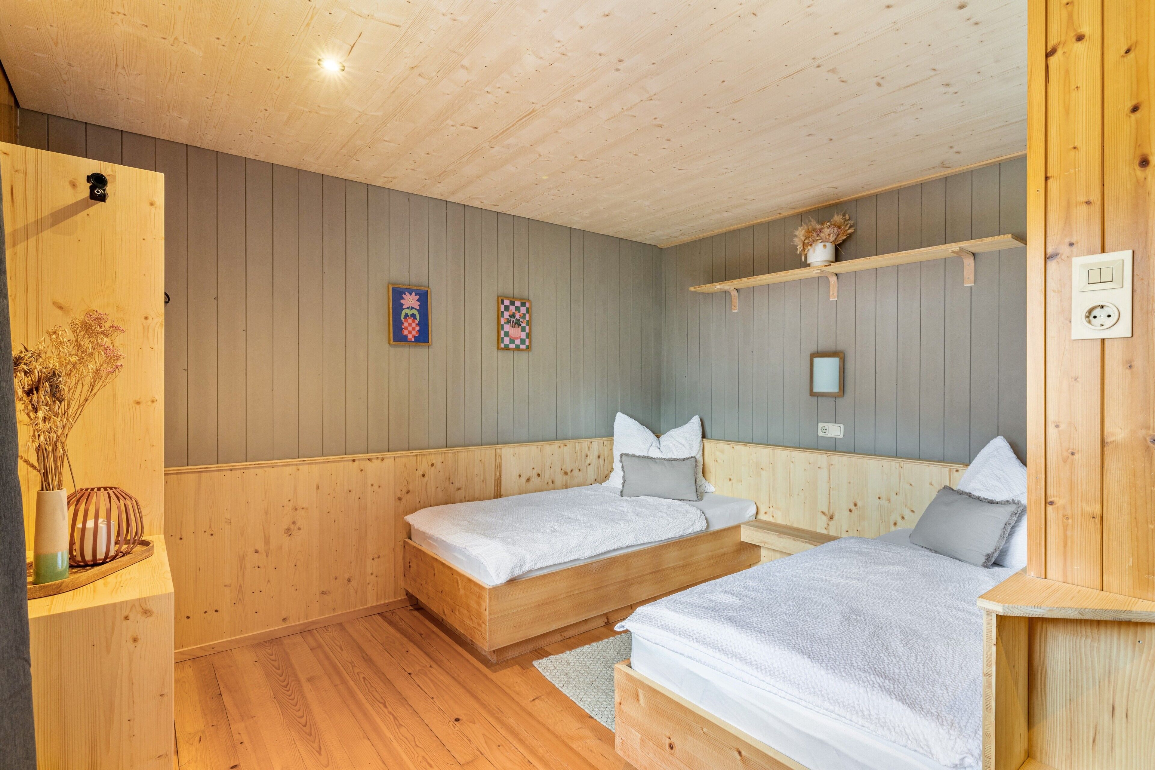 Una habitación con dos camas de madera, un estante con una planta y una lámpara fijada a la pared.