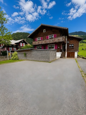 A two-story wooden building with a balcony, a paved driveway, and a mountain backdrop.