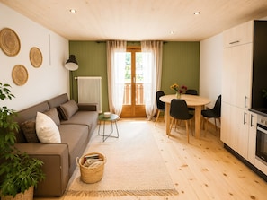 A cozy living room with a sofa, a dining table with chairs, a kitchen area with cabinets and a stove, and a large window with curtains.