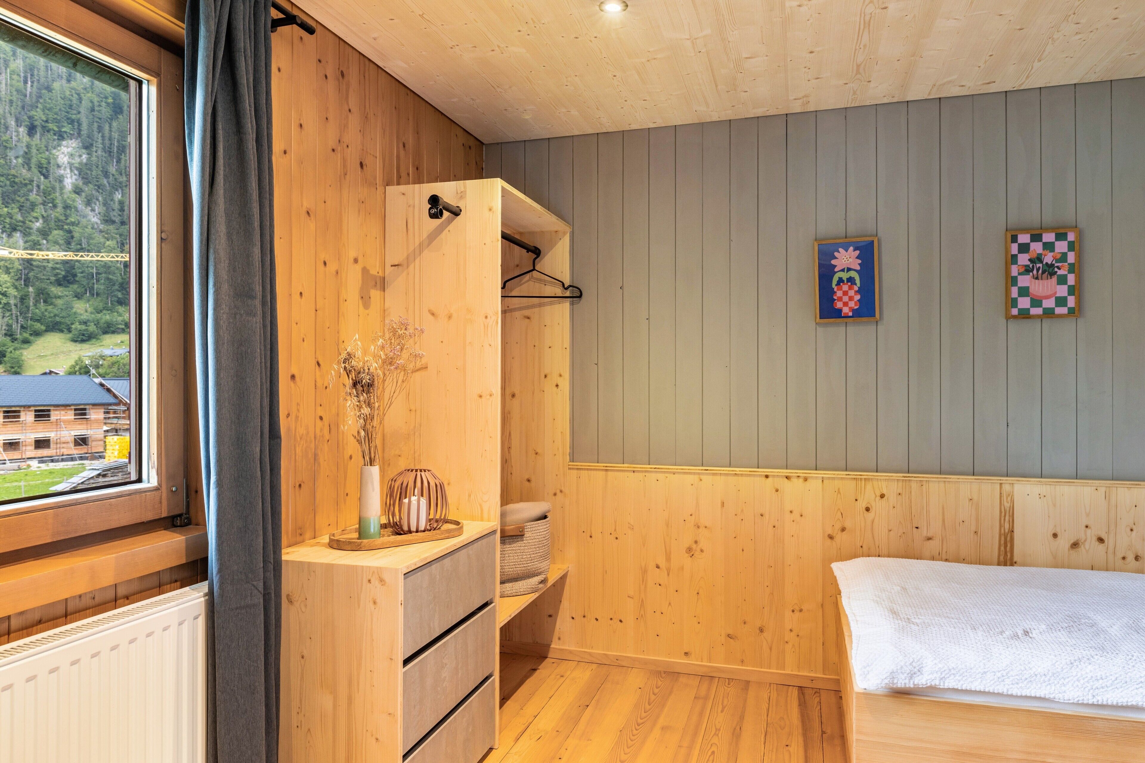Una habitación con paredes de madera, una cama, un aparador, un ventanal con vista a un edificio y un estante de pared con un jarrón.
