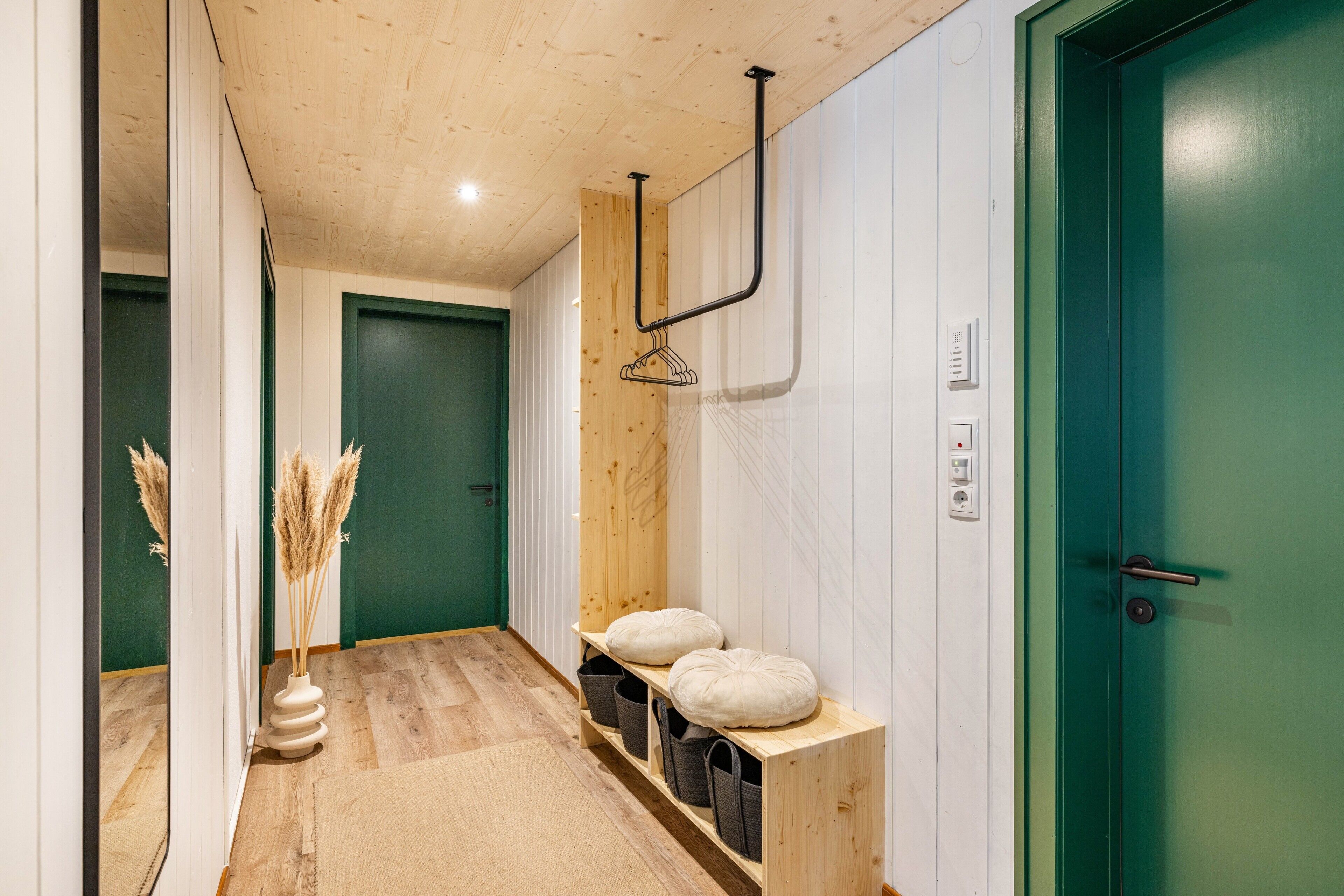 Ein Korridor mit Holzboden, einer grünen Tür, einer weißen Wand mit einem angebauten Kleiderhaken und einem kleinen Regal mit Kissen und Körben.