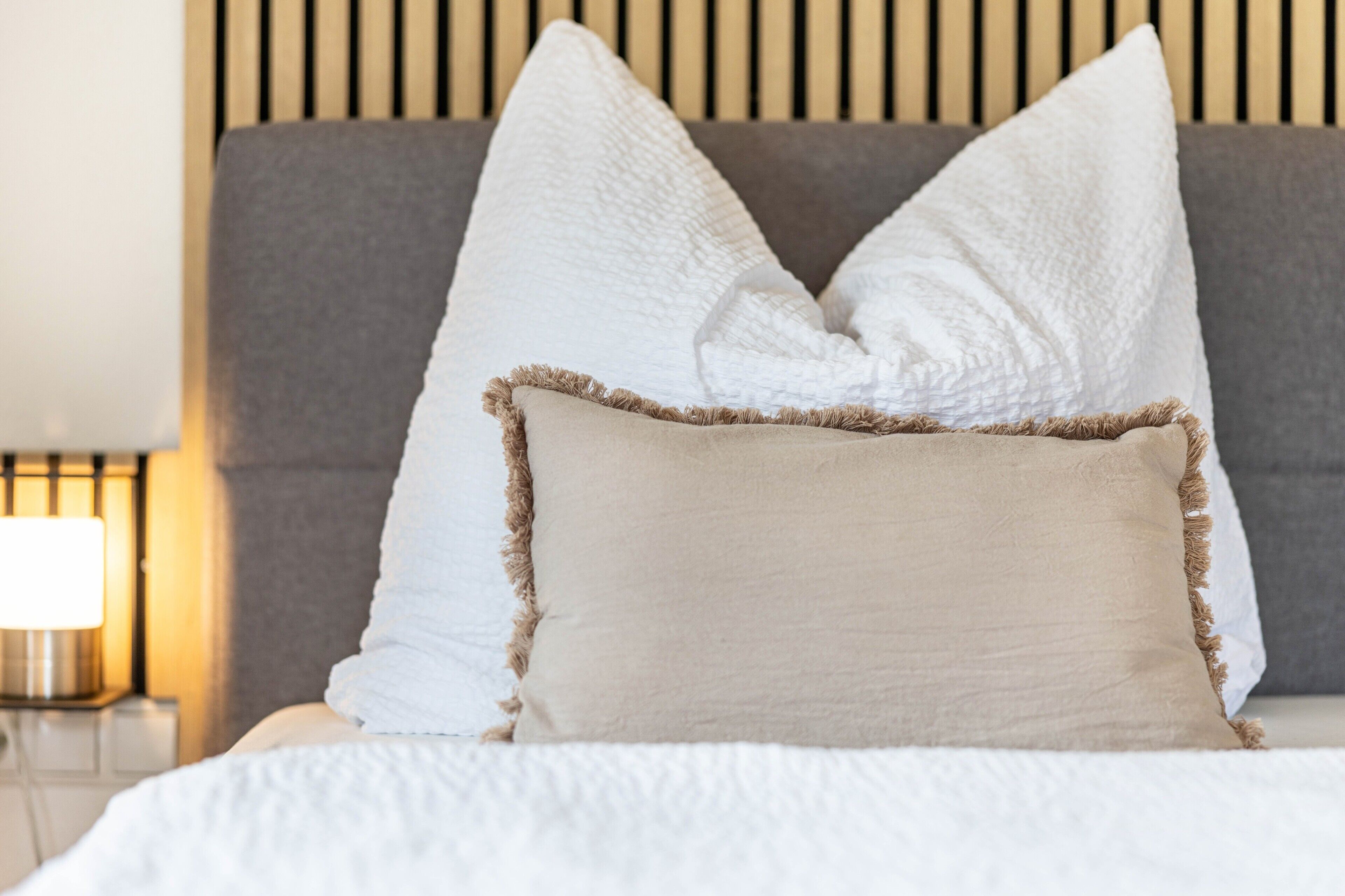 Un lit avec des oreillers blancs et beige, une tête de lit grise et une lampe de chevet.