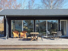 Un edificio moderno con fachada oscura, amplios ventanales de vidrio y una terraza de madera con mobiliario exterior.