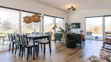 A spacious living area with a dining table, chairs, and a sofa. There are large windows providing natural light and a view of the outdoors.