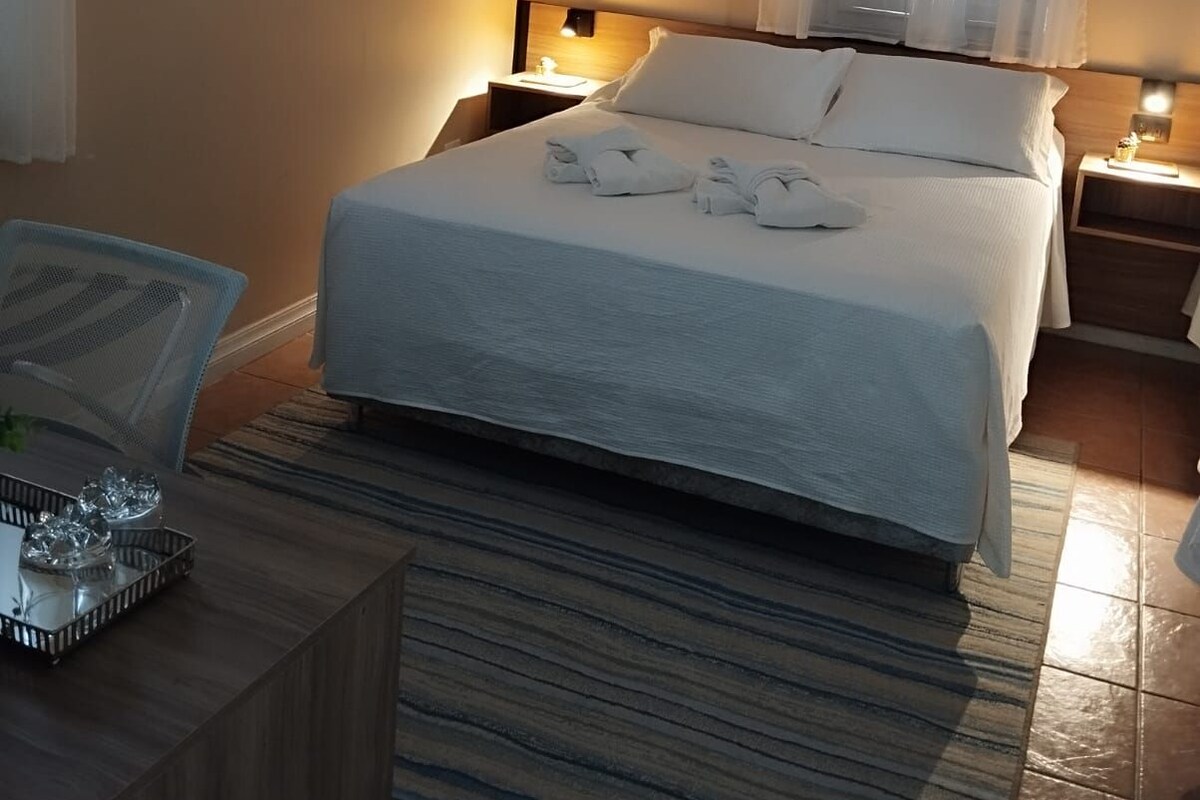 Een hotelkamer met een bed, nachtkastje, lamp, raam met gordijnen en een airconditioning aan de muur.