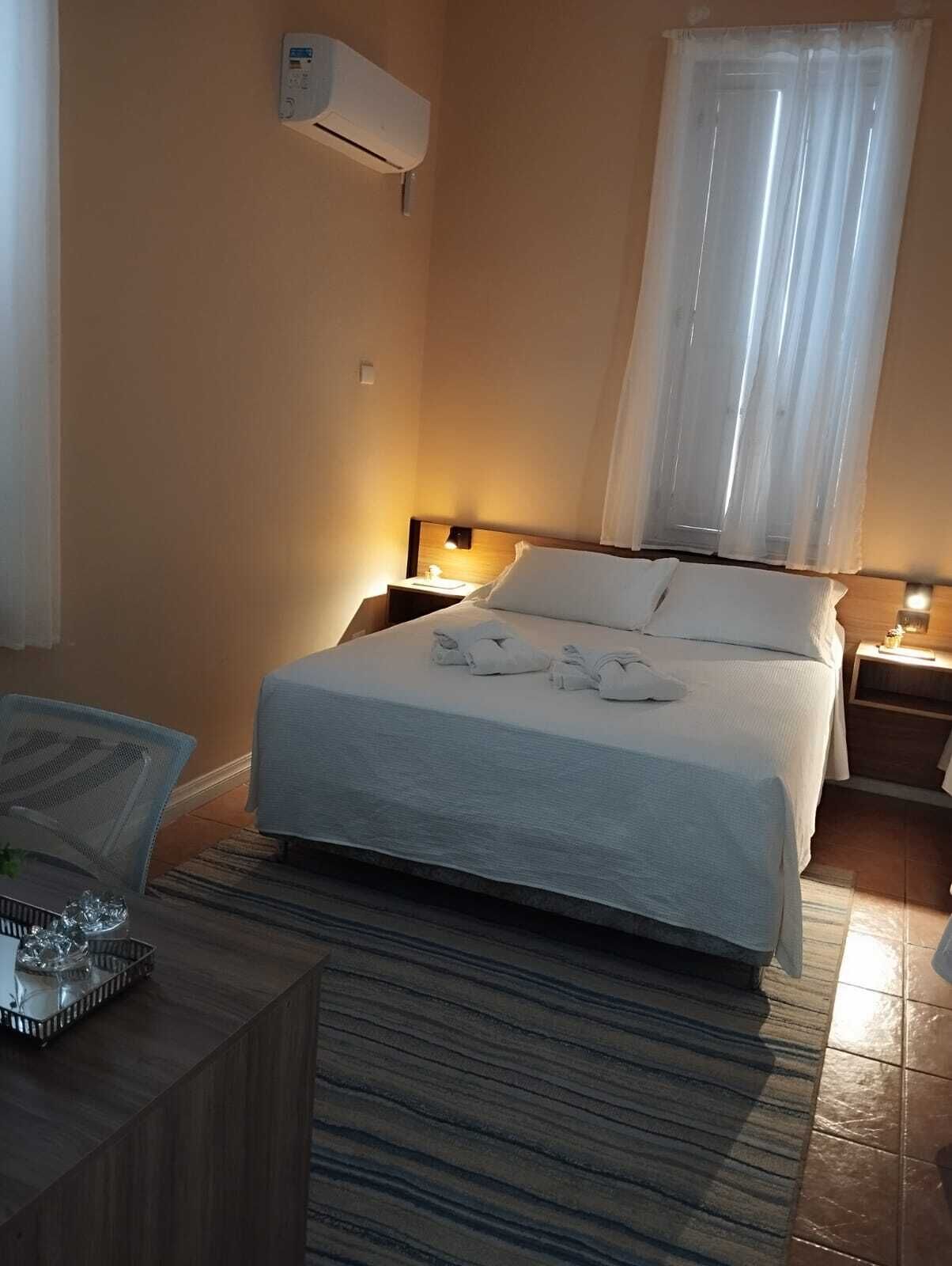 Une chambre d’hôtel comprenant un lit, une table de chevet, une lampe, une fenêtre avec des rideaux et un appareil de climatisation.