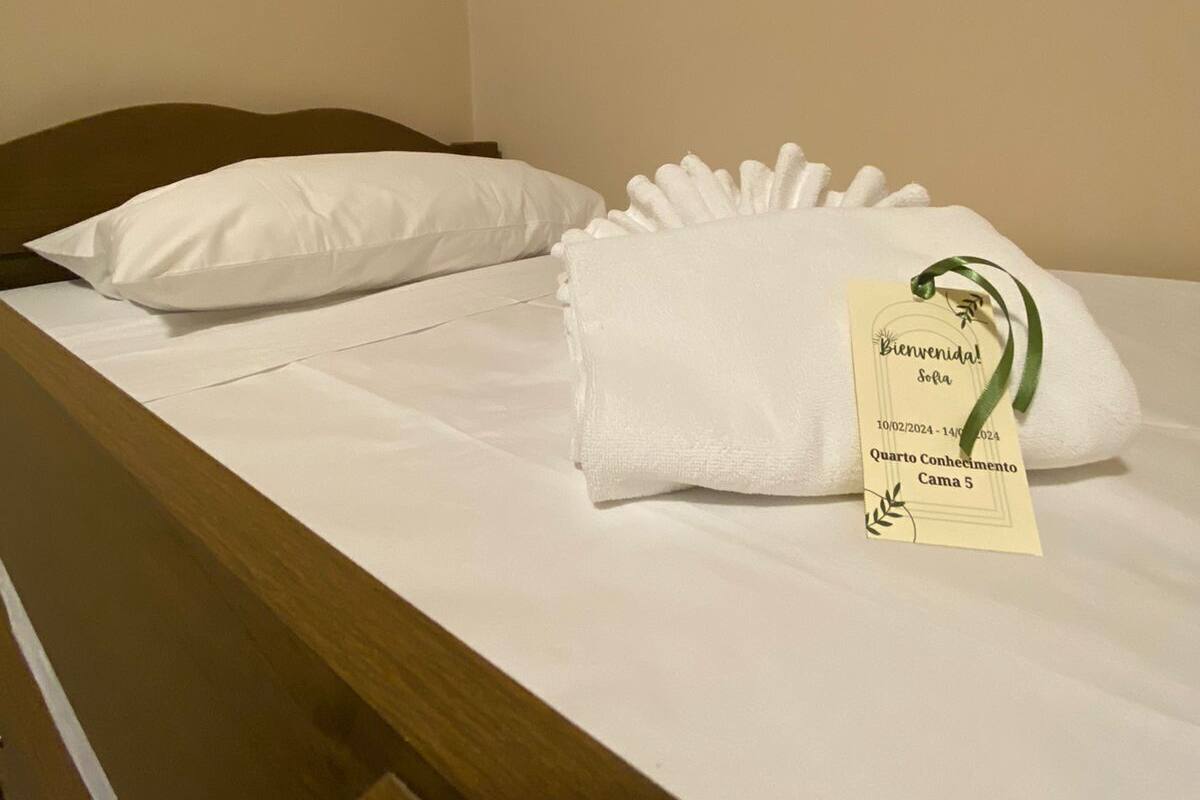 A neatly made bed with white linens and pillows, a tag indicating a stay from 10/02/2024 to 14/02/2024, and a label reading "Quarto Conhecimento Cama 5".