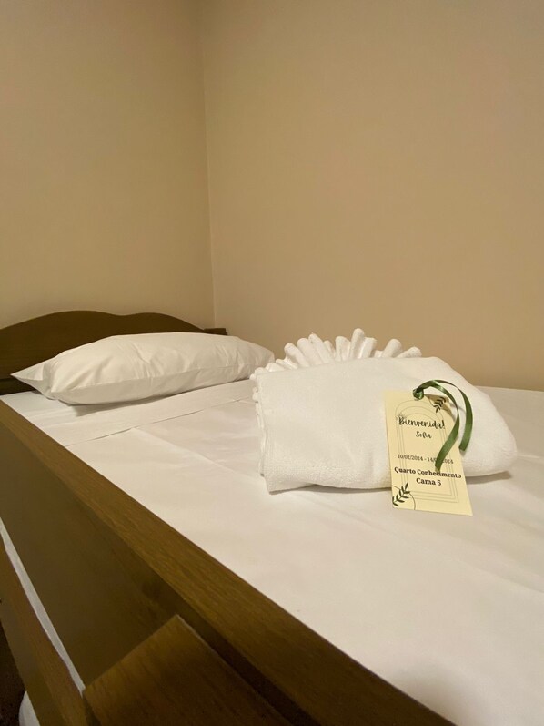 Un lit bien fait, avec du linge de lit blanc et des oreillers, une étiquette indiquant une période de séjour du 10/02/2024 au 14/02/2024, et une étiquette portant l’inscription « Quarto Conhecimento Cama 5 ».